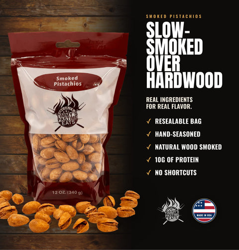 A bag of Smoke Paste Smoked Pistachios displayed on a dark wood background with scattered whole and cracked pistachios in front. The packaging is red, white, and clear, showing the pistachios inside.